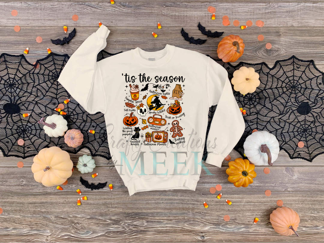 White sweatshirt with Halloween-themed design on a wooden surface with pumpkins and spider webs.